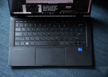 The HP Elite Dragonfly Max keyboard seen from above on a fabric bench. The screen displays The Verge homepage.