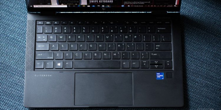 The HP Elite Dragonfly Max keyboard seen from above on a fabric bench. The screen displays The Verge homepage.