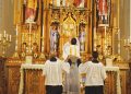 The Rev. Trenton Rauck elevates a chalice for consecration during a Latin Mass at Sacred Heart Catholic Church as servers lift his chasuble vestment. - PHOTO BY SCOTT REEDER
