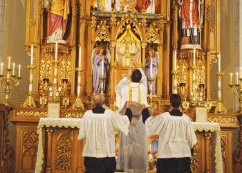 The Rev. Trenton Rauck elevates a chalice for consecration during a Latin Mass at Sacred Heart Catholic Church as servers lift his chasuble vestment. - PHOTO BY SCOTT REEDER