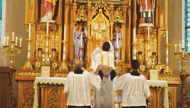 The Rev. Trenton Rauck elevates a chalice for consecration during a Latin Mass at Sacred Heart Catholic Church as servers lift his chasuble vestment. - PHOTO BY SCOTT REEDER