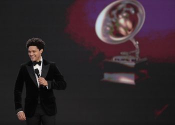 A man in a tuxedo holding a microphone onstage. A Grammy trophy is projected behind him.