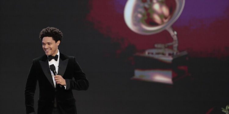 A man in a tuxedo holding a microphone onstage. A Grammy trophy is projected behind him.