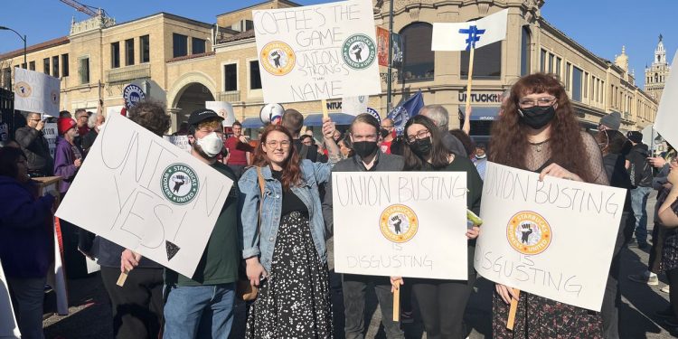 Starbucks workers stand in the street holding signs that read, “Union yes,” “Union busting is disgusting,” and “Coffee’s the game, union strong’s the name.”