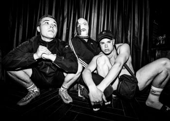 Three band members, one with a knit cap over his face, crouch down.