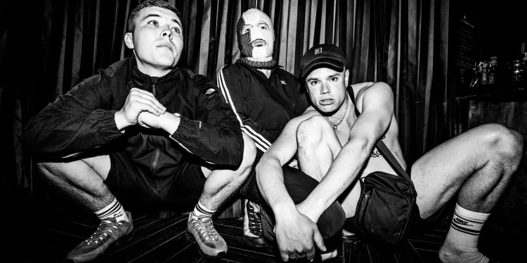 Three band members, one with a knit cap over his face, crouch down.