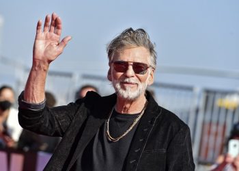 A man with gray hair and a beard waves as he walks.