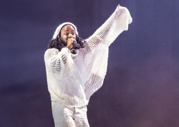 A man in a woven white shirt holds up one arm and sings into a hand-held microphone.