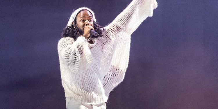 A man in a woven white shirt holds up one arm and sings into a hand-held microphone.
