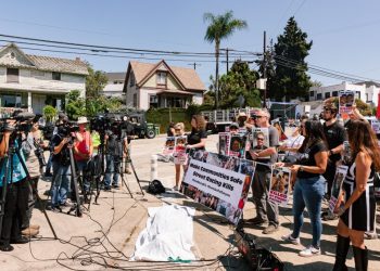 TV cameras pointed at a crowd of people with protest signs and banners, standing behind a white sheet covering a fake body