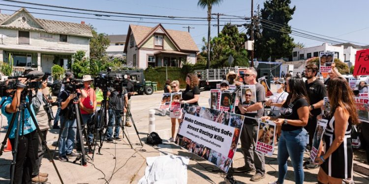 TV cameras pointed at a crowd of people with protest signs and banners, standing behind a white sheet covering a fake body