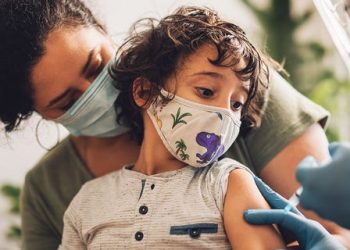 A child in a mask, held by a parent, is shown receiving a vaccine. 
