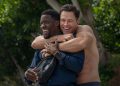 A shirtless man hugs another man from behind.