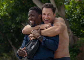 A shirtless man hugs another man from behind.