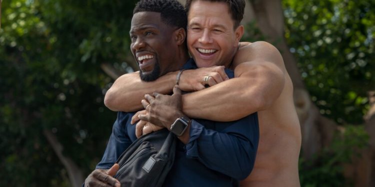 A shirtless man hugs another man from behind.
