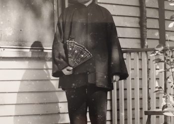 An old black-and-white photo of a man standing on a porch, a small dog at his feet.