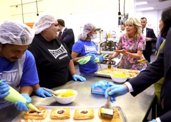First Lady Jill Biden speaks with people who are putting glaze on pastries at a bakery table.
